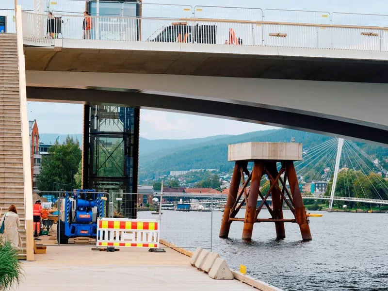 En brygge med en ny, stålbru over med heis. Og en elv som renner på siden med et stort brufundament i elva.