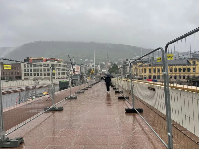 Fortau i rød granittstein over en bru som holder på å bli bygd, med anleggsgjerder på begge sider. Noen mennesker går i retning byen med åskam bak i regnvær og tåke (foto)