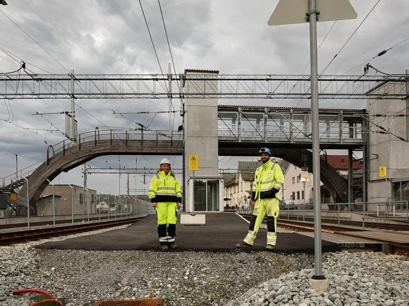 To smilende mennesker i gult verneutstyr og hjelm. Står på Moss togstasjon som er under utbygging. Overgangsbru i tre i bakgrunn.