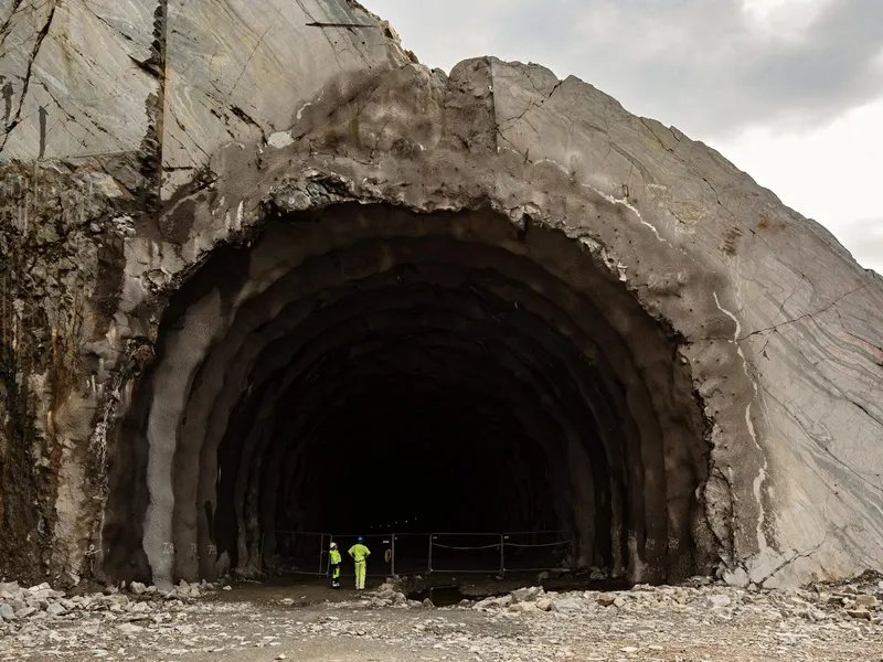 Bildet viser to anleggsarbeidere i gult verneutstyr som står foran en stor, svart tunnel.