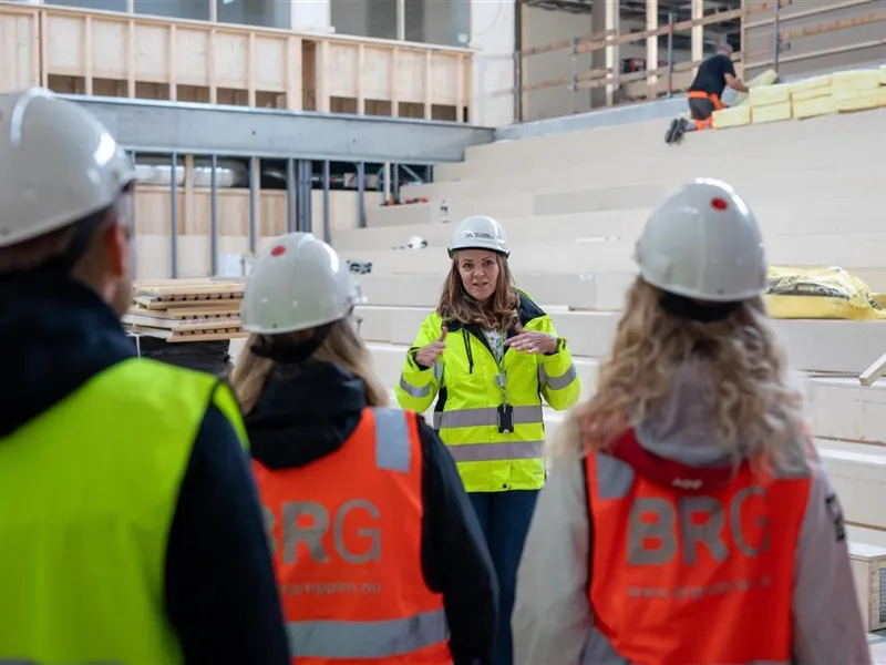 En gruppe med lærere og prosjektleder i refleksvester og hjelmer står samlet inne i et rom på byggeplassen under en omvisning. - Klikk for stort bilde