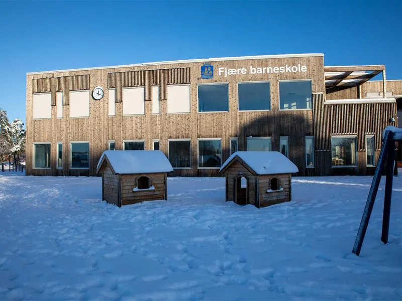 Fasaden til Fjære barneskole en vinterdag, med trepanel, store vinduer og et lite lekehus i snøen foran bygget. - Klikk for stort bilde