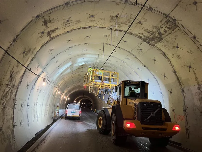 I en tunnel står en gravemaskin med lift. To personer i refleksklær monterer noe i tunneltaket.   - Klikk for stort bilde