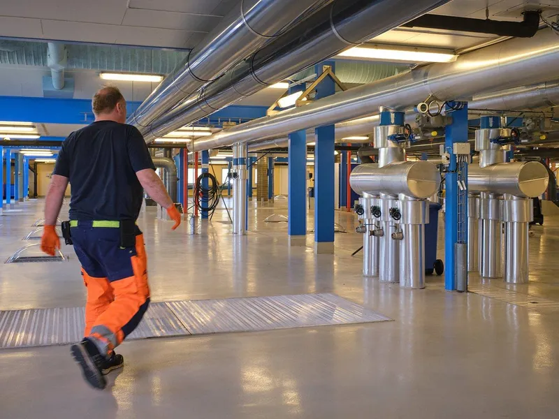 Mann i oransje arbeidsbukse går vekk fra kamera med ryggen til inne på et renseanlegg. Store stålrør til høyre i bildet.