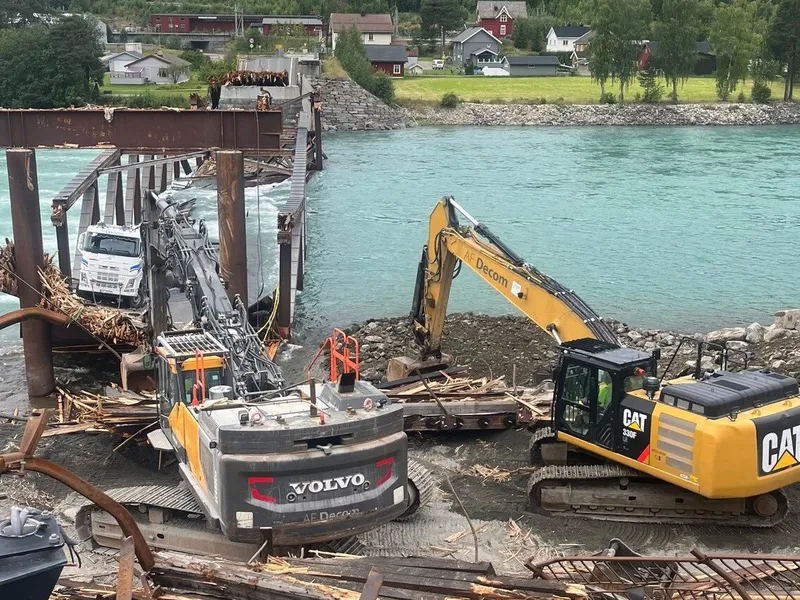 To gravemaskiner samt en lastebil river broen over vannet. Rester av planker og avkapp. 