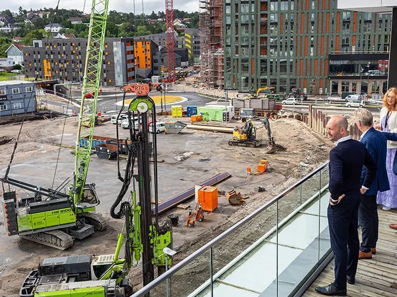 Tre menn og en dame står på en veranda og ser utover en byggeplass. - Klikk for stort bilde