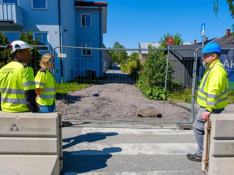 en dame og to menn i gule vester foran en vei som skal oppgraderes.