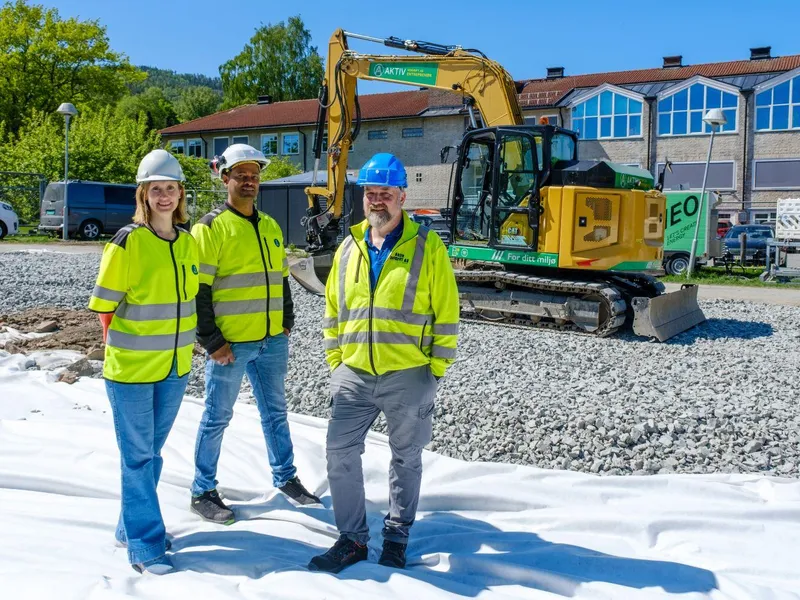 en dame og to menn foran en elektrisk gravemaskin. 