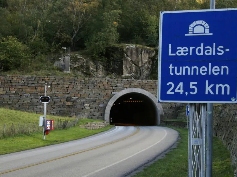 Eit skilt ved inngangen til Lærdalstunnelen viser at tunnelen er 24,5 km lang, med ein veg som fører inn i tunnelen omgitt av natur.