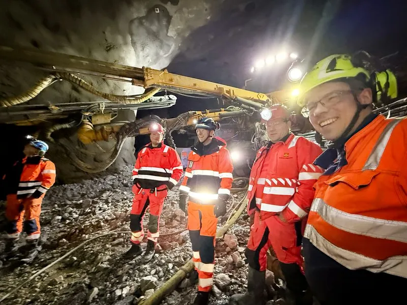 Bildet er tatt inne i tunnelen med en gul fjellboremaskin i brakgrunnen og i skinnet fra hodelykert og maskinens arbeidslys. Bildetektsen sier, Skanska passerer 15 000 meter drevet tunnel på OPS Hålogalandsvegen.  I marmorhvelvet i Gullberg øst, som vender ut mot Tjeldsundbroen står fra venstre: Statens vegvesen ved Bjørn-Einar Hansen, Hålogalandsvegen AS ved Kent Gøran Johansen, Statens vegvesen ved Henning Larsen og sprengningsbas Tore Gunnar Gundersen.