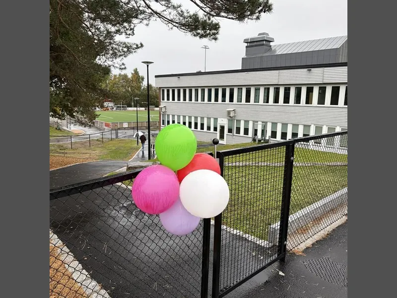 Ballonger på en port inn til en barnehage. Bak ser vi barnehagen, en asfaltert gangvei og en kunstgressbane - Klikk for stort bilde