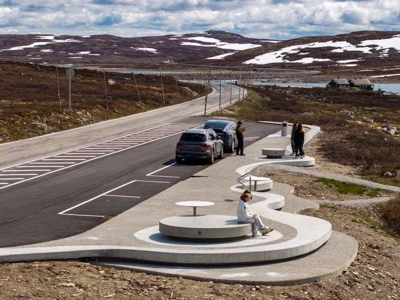 To ungjenter sit på runde rastemøblar i betong og ser på utsikta over innsjøen Ørteren på Hardangervidda. Nokre turistar tek bilete av kvarandre i bakgrunnen.