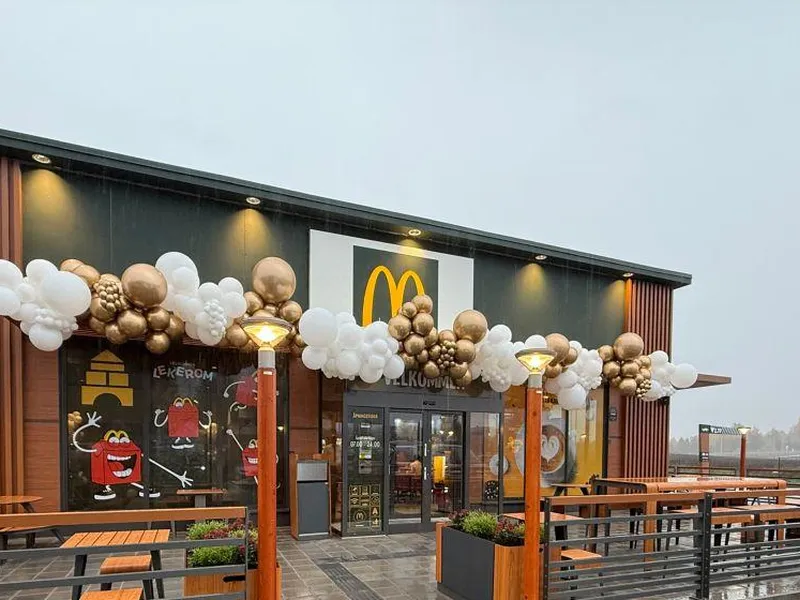 Utsiden av en McDonald's-restaurant med trepanel og store vinduer. Over inngangspartiet henger det dekorasjoner av hvite og gullfargede ballonger. Det er utemøbler i form av trebenker og bord, med rekkverk som omgir uteserveringsområdet. På vinduene er det klistremerker med McDonald's-karakterer, inkludert Happy Meal-figurer. Parkeringen foran restauranten er våt, noe som tyder på at det nylig har regnet, og i bakgrunnen kan man se et vinterlig landskap. Foto.