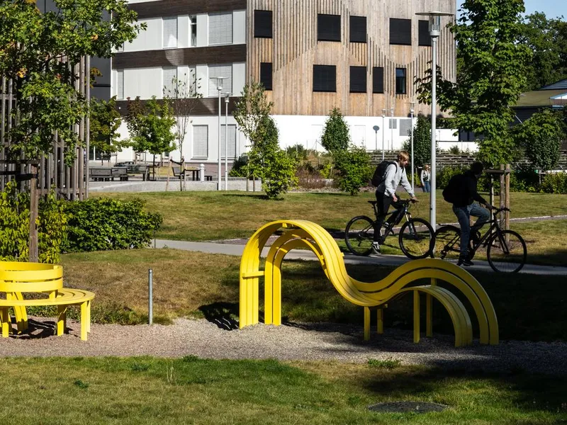 Grønt område utenfor skole. Gule dekorative sittebenker som også fungerer som lekestativ. To syklister. Foto
