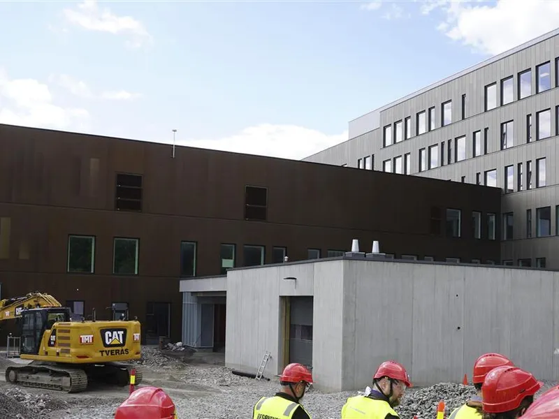 Bilde av grå og brun bygning i bakgrunnen med firkantet betongpåbygg i forkant, syv personer med hjelm og refleksvest går i forkant av byggeplassen - Klikk for stort bilde