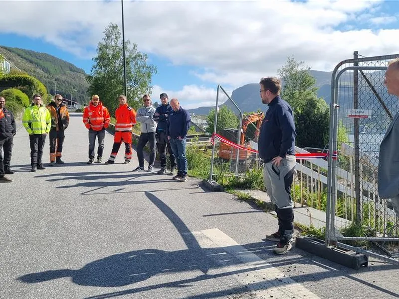 Personar ikledd arbeidsklede ventar på at ordførar skal klippe snora for bygginga - Klikk for stort bilete