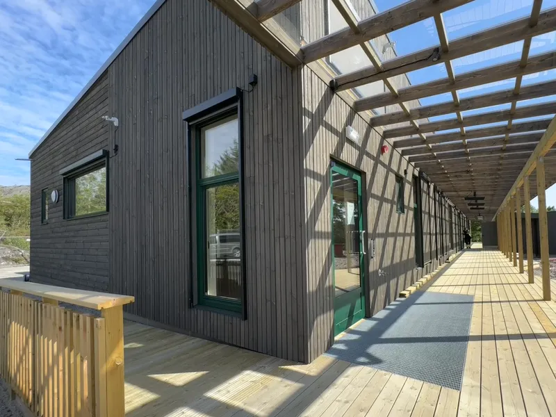 A modern wooden building with large windows, featuring a covered walkway and a wooden deck. Clear blue sky in the background.