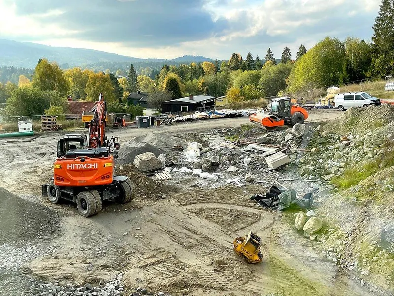 Bildet viser et anleggsområde, med blant annet gravemaskin og vibrovalse. I bakgrunnen ses bolighus.