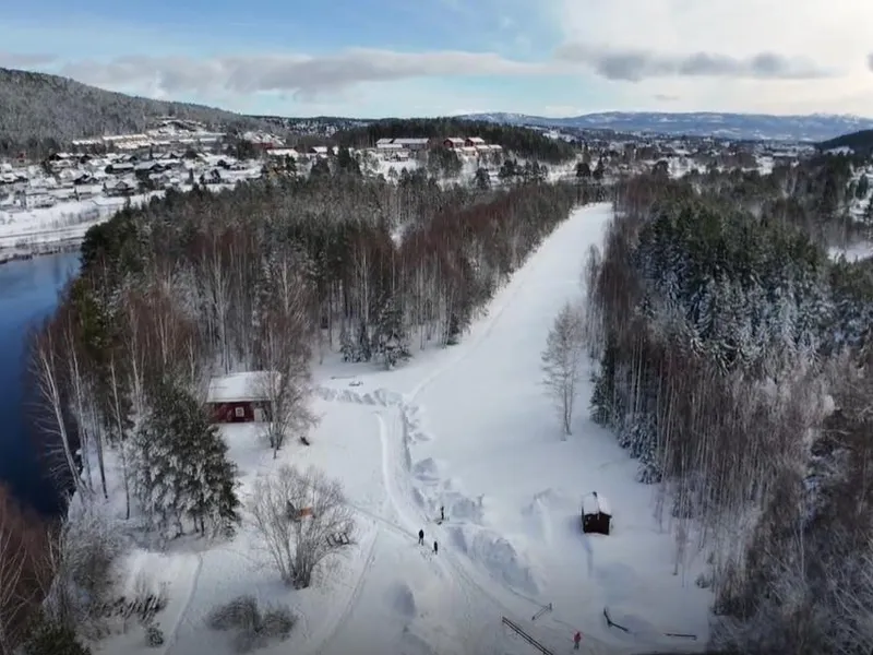 Flyfoto av et snødekt landskap med en elv, skog og noen bygninger i bakgrunnen.