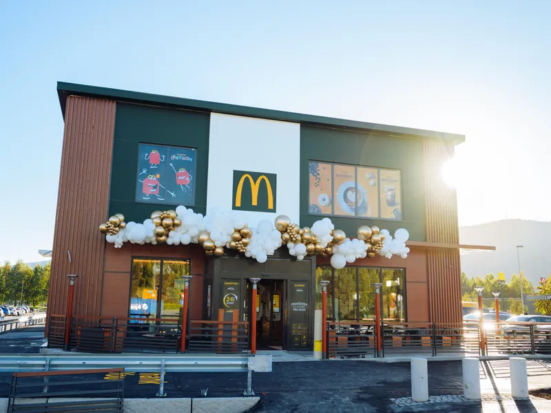 Fasaden på en McDonald's-restaurant, dekorert med ballonger i gull og hvitt for en spesiell anledning. Det ikoniske gule McDonald's-logoen er synlig på bygningen, mens morgensolen skinner i bakgrunnen. Foto.