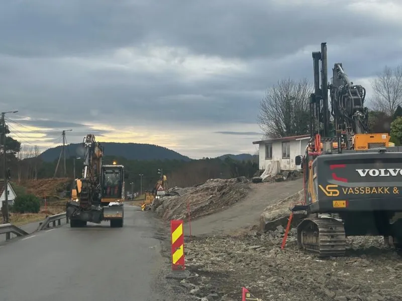 To gravemaskiner er i arbeid langs ein veg med ein garasje i bakgrunnen.
