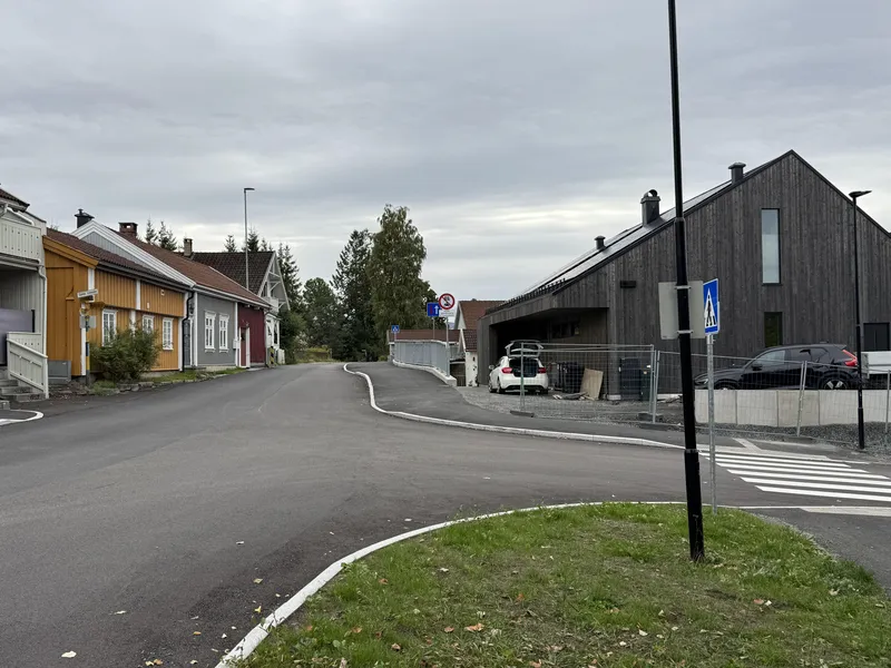 En stille gate med ulike bygninger, inkludert fargerike trehus på venstre side og en moderne, grå bygning til høyre, under en overskyet himmel.