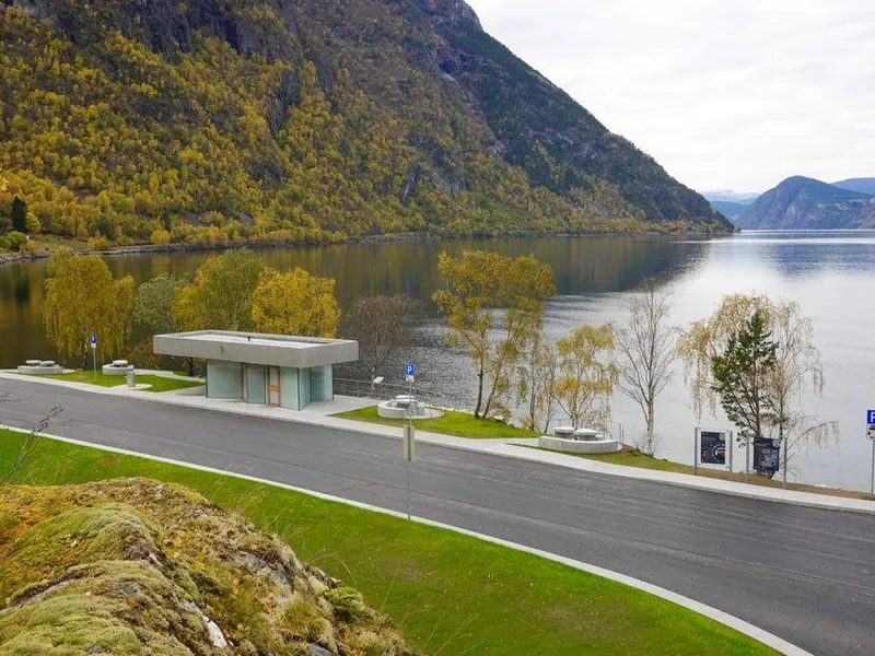 Oversiktsbilete av rasteplassen på Røyrnes. Eit toalettbygg ligg mellom vegen og fjorden, omgjeve av sitjeplassar, tre og bratte fjellsider med haustfargar.