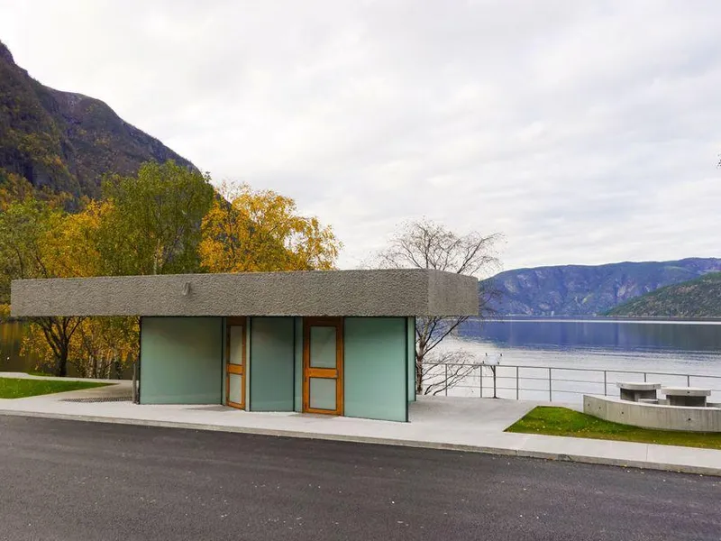 Toalettbygg i betong og glas på Røyrnes ved Lærdalsfjorden. Framfor bygget ligg asfalt og gangareal, medan fjorden, fjell og haustfarga tre dannar bakgrunnen.