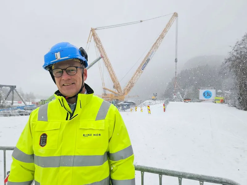 Portrettbilde av mann i gul arbeidsjakke og hvit hjelm foran stor gul heisekran.