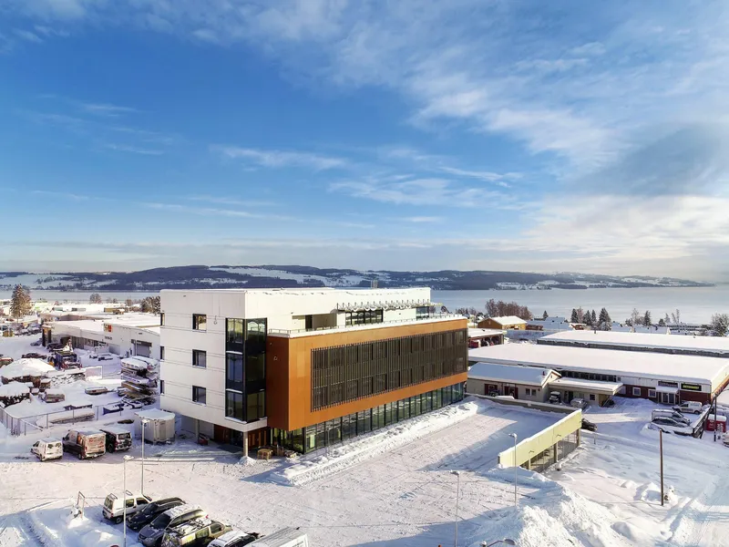 Foto av kontorbygg med en hvit del og en mørkere del, vinterlandskap med Mjøsa i bakgrunnen