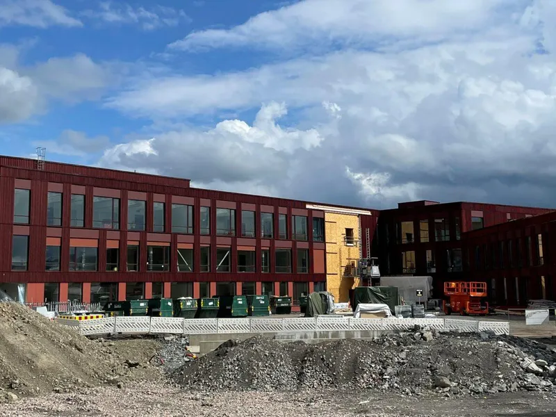 Byggeplass, utvendig, langstrakt bygg o 2-3 etasjer, rød fasade, blåp himmel, byggegrop i forgrunnen