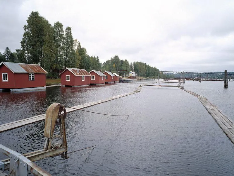 En rekke røde naust av tre langs kanten av en rolig innsjø, med trær i bakgrunnen og en trebrygge som strekker seg ut i vannet under en overskyet himmel.