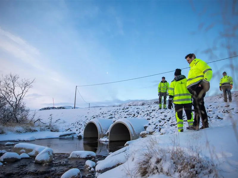 Flere mennesker i varselgule vinterklær står ved en elv og et par nye stikkrenner. Bildet er tatt på vinterstid. Det er klar, blå himmel. - Klikk for stort bilde