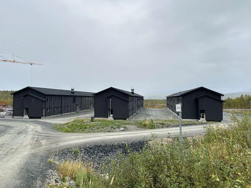 Bygninger med mørk fasade i Camp Akkasæter omgitt av grå veier og vegetasjon. En kran står i bakgrunnen.