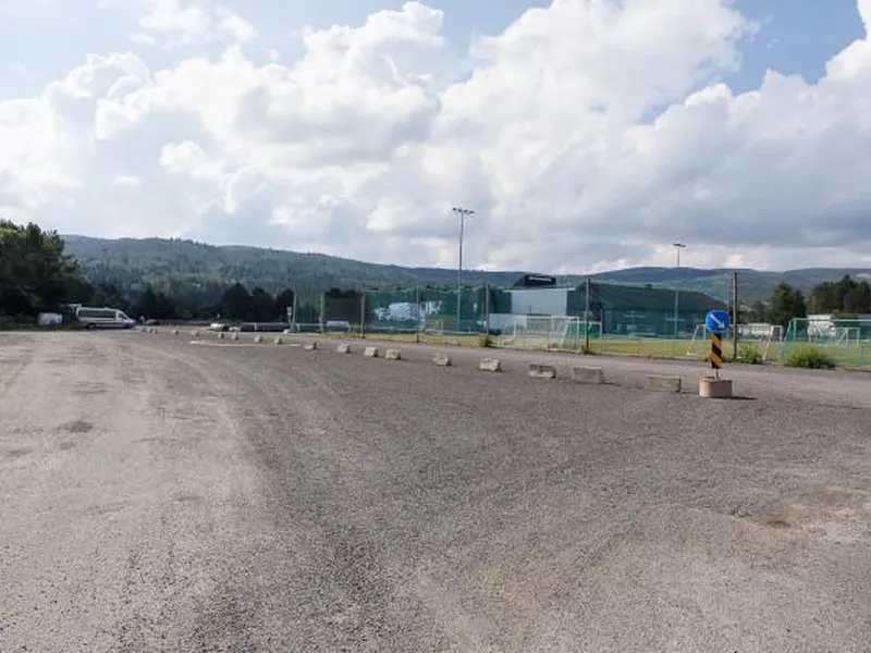 Parkeringsplass i grus med Geilohallen i bakgrunnen. Bilde. 