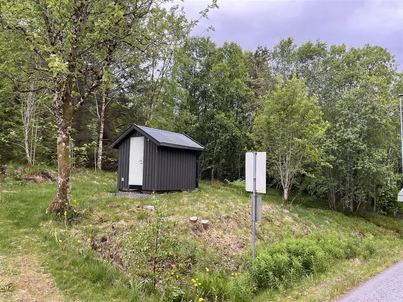 Lita bygning i skogkanten ved ein veg. - Klikk for stort bilete