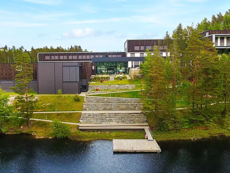 Bilde av skolen fra utsiden. Vann på siden og skog rundt. 