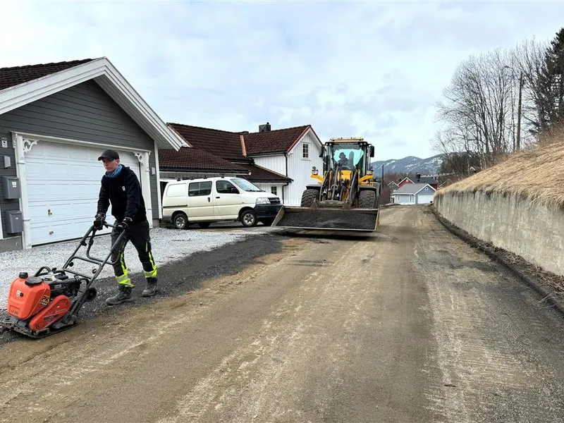 Mann med hoppetusse foran anleggsmaskin i ei bygate. Foto - Klikk for stort bilde