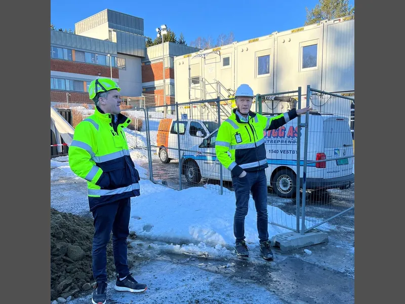 To menn i vernejakke og hjelm på byggeplass. Den ene peker og forklarer. - Klikk for stort bilde