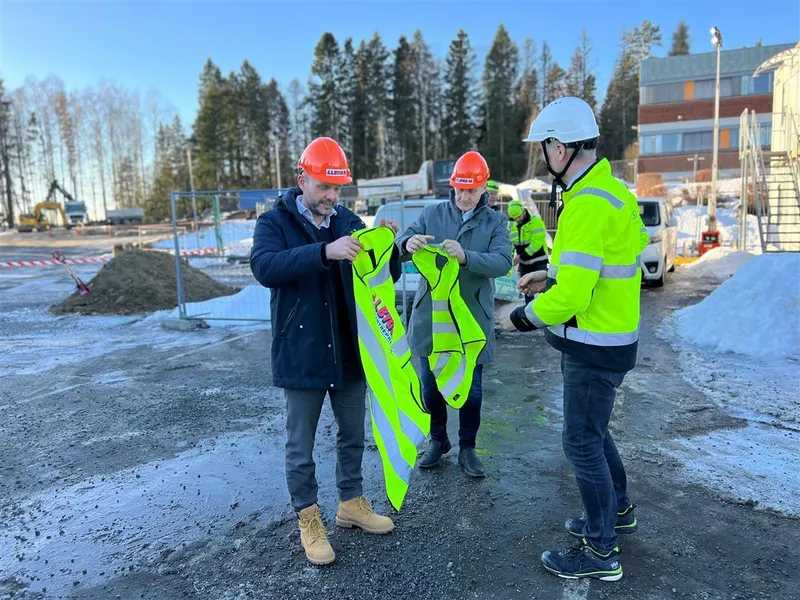 Mann i verneutstyr deler ut refleksvest og hjelm til to andre menn på byggeplass.  - Klikk for stort bilde