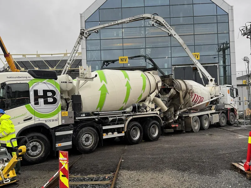 To betongbiler med betongpumper foran eksisterende toghall. Et jernbanespor i forgrunnen.
