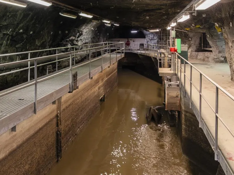 En underjordisk tunnel med vann langs midten og gangvei med rekkverk på begge sider.