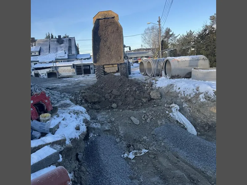 En dumper har tømt jord på anleggsområdet, med store betongrør stablet langs kanten og snø rundt. - Klikk for stort bilde